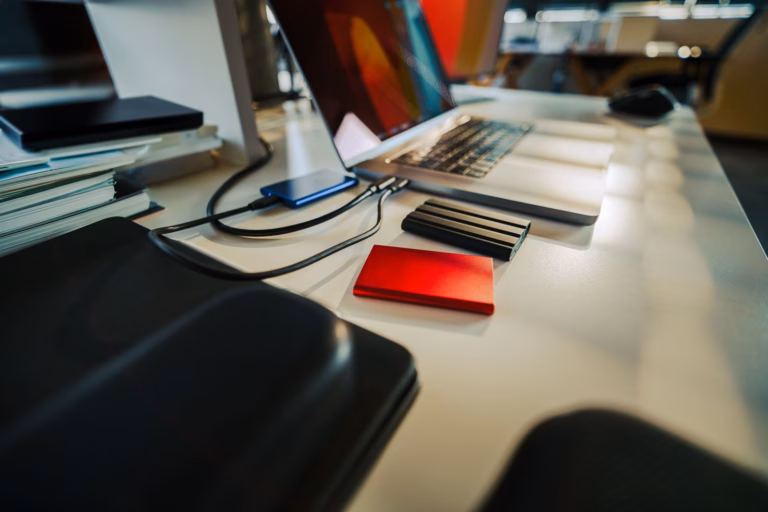 An open laptop on a desk beside a cable-connected portable SSD, a stack of external backup drives and a pile of paper notebooks.