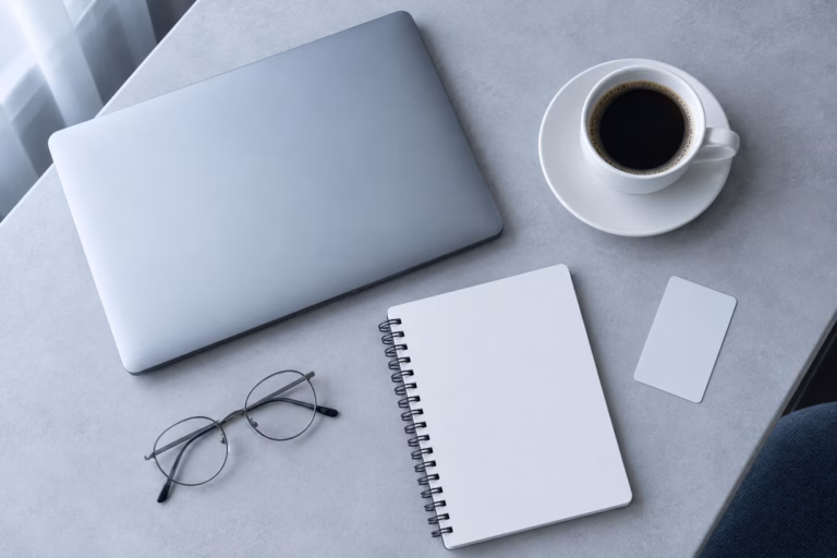 Top-down editorial flat lay of a hotel desk: a closed silver laptop, a white coffee cup, a blank notebook, an unmarked key card and thin reading glasses.