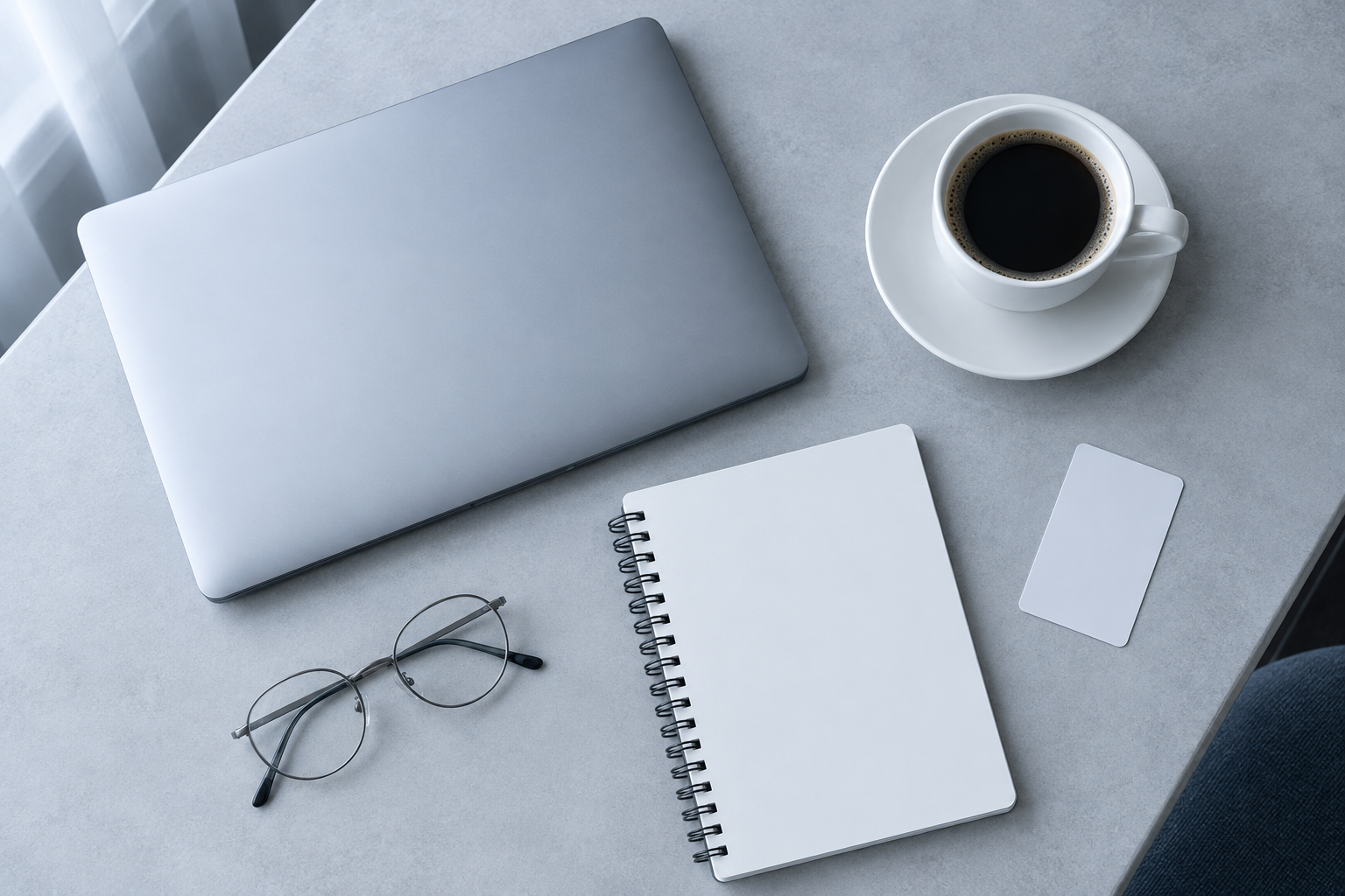 Top-down editorial flat lay of a hotel desk: a closed silver laptop, a white coffee cup, a blank notebook, an unmarked key card and thin reading glasses.
