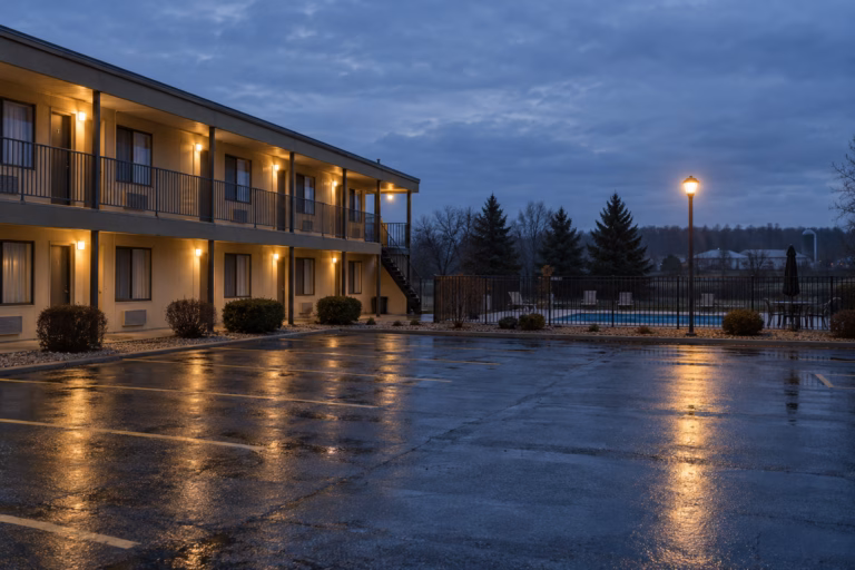 A modest two-story roadside motel exterior at dusk, warm lamplight on a walkway, an empty rain-wet parking lot in front, and a fenced pool area under a pale overcast sky