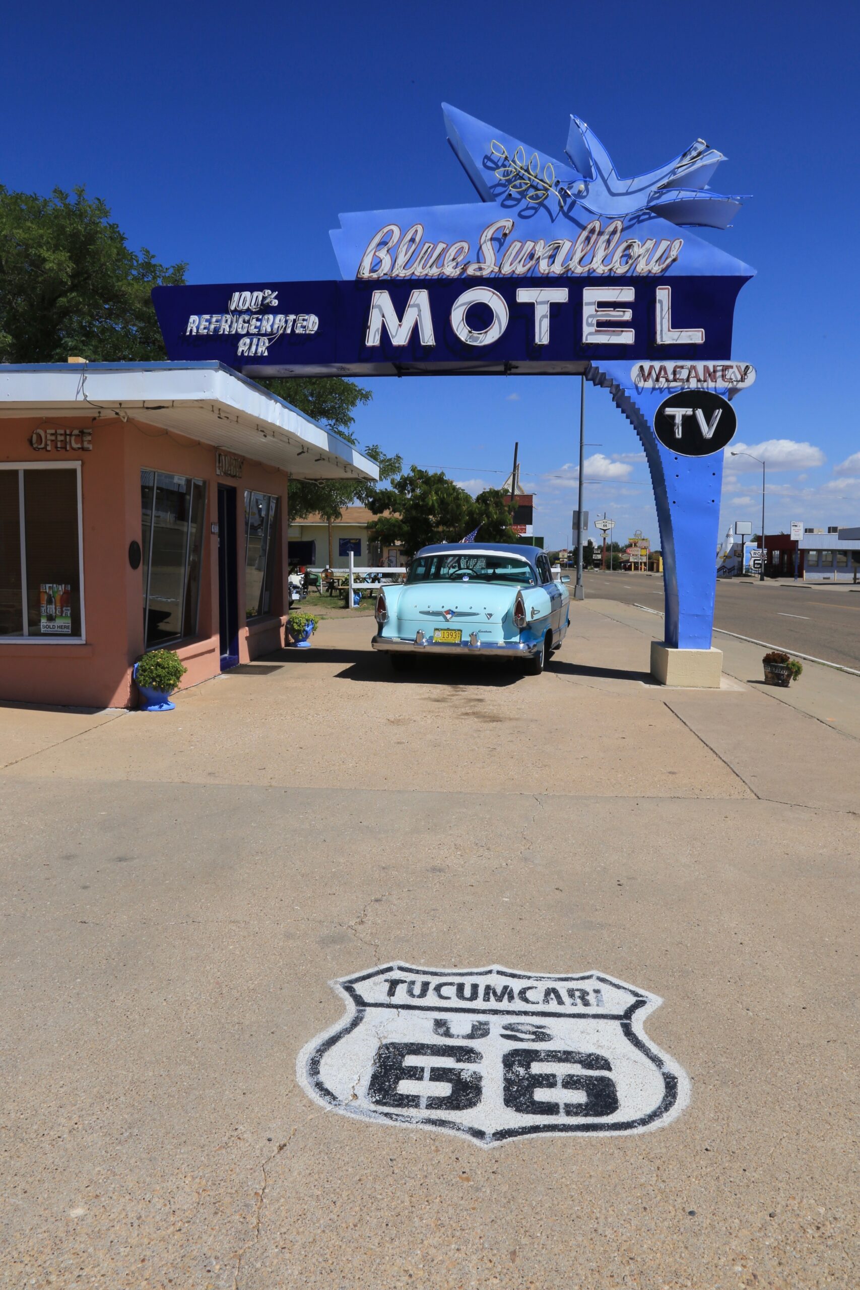 A vintage scene featuring the iconic Blue Swallow Motel sign and a classic car parked beneath it, with a Route 66 marker on the ground, under a clear blue sky.