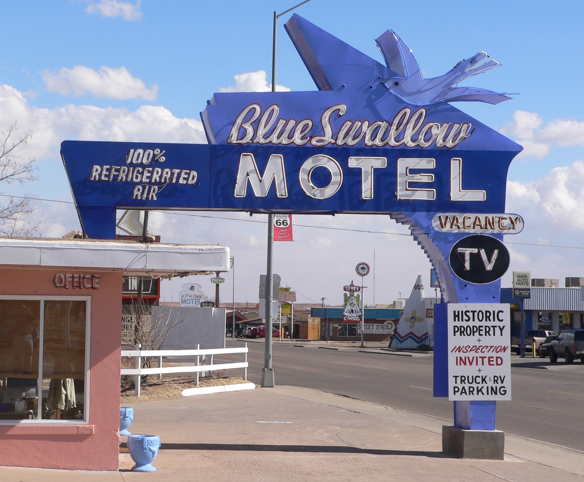 A vibrant blue sign for the Blue Swallow Motel, featuring retro design elements and a large bird silhouette, with a clear view of the motel's entrance and a 'TV