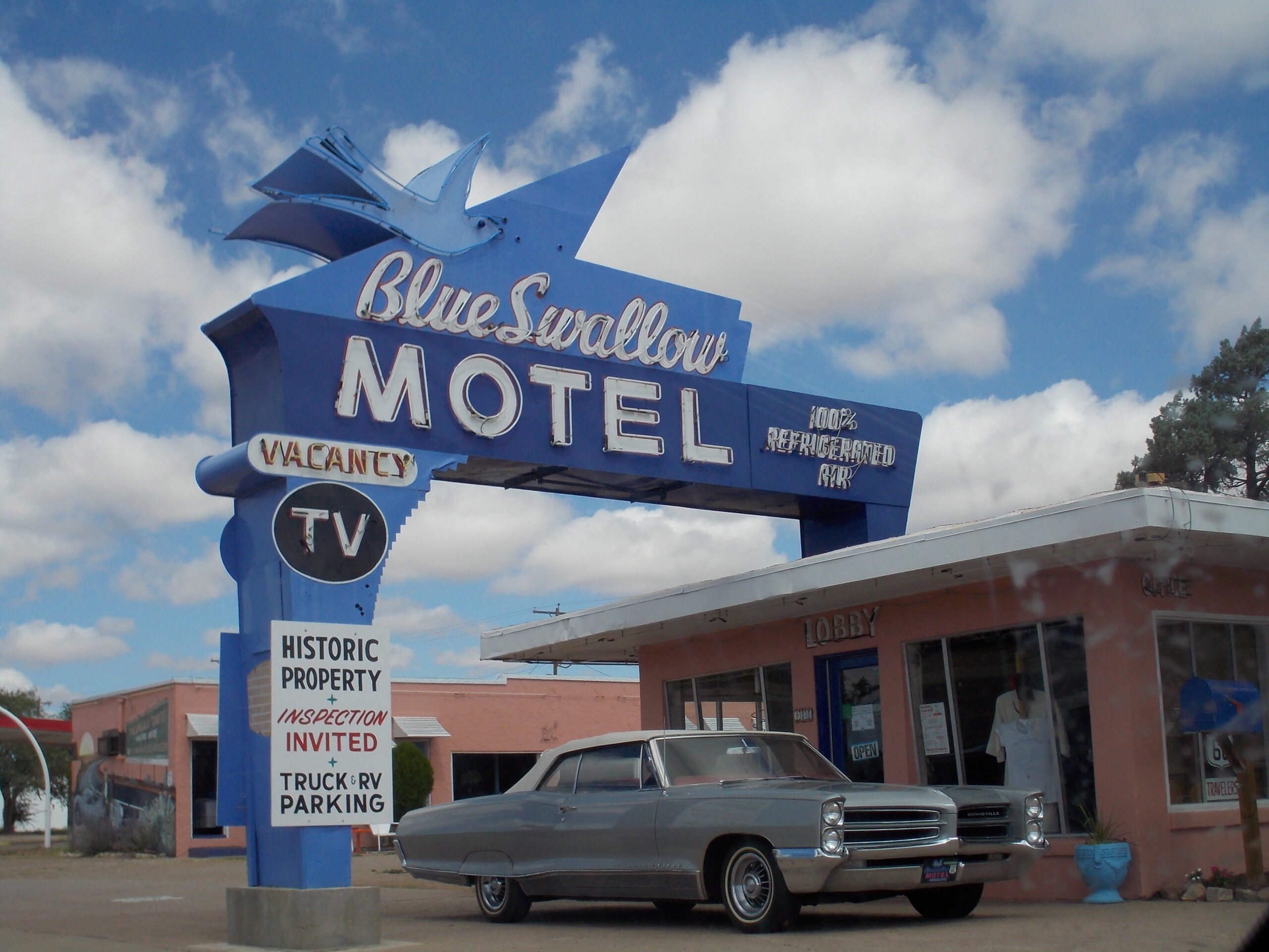 A vintage sign for the Blue Swallow Motel with a retro car parked in front, under a clear sky with clouds. The motel's pink building and lobby entrance are also