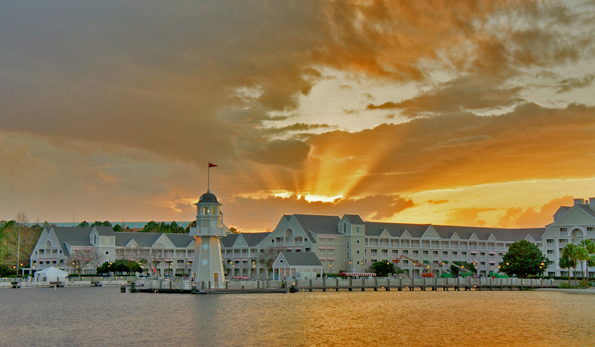 Sunset at Disney's Yacht Club Resort with a lighthouse and dock in the foreground, featuring the resort's distinctive architecture and a dramatic sky.