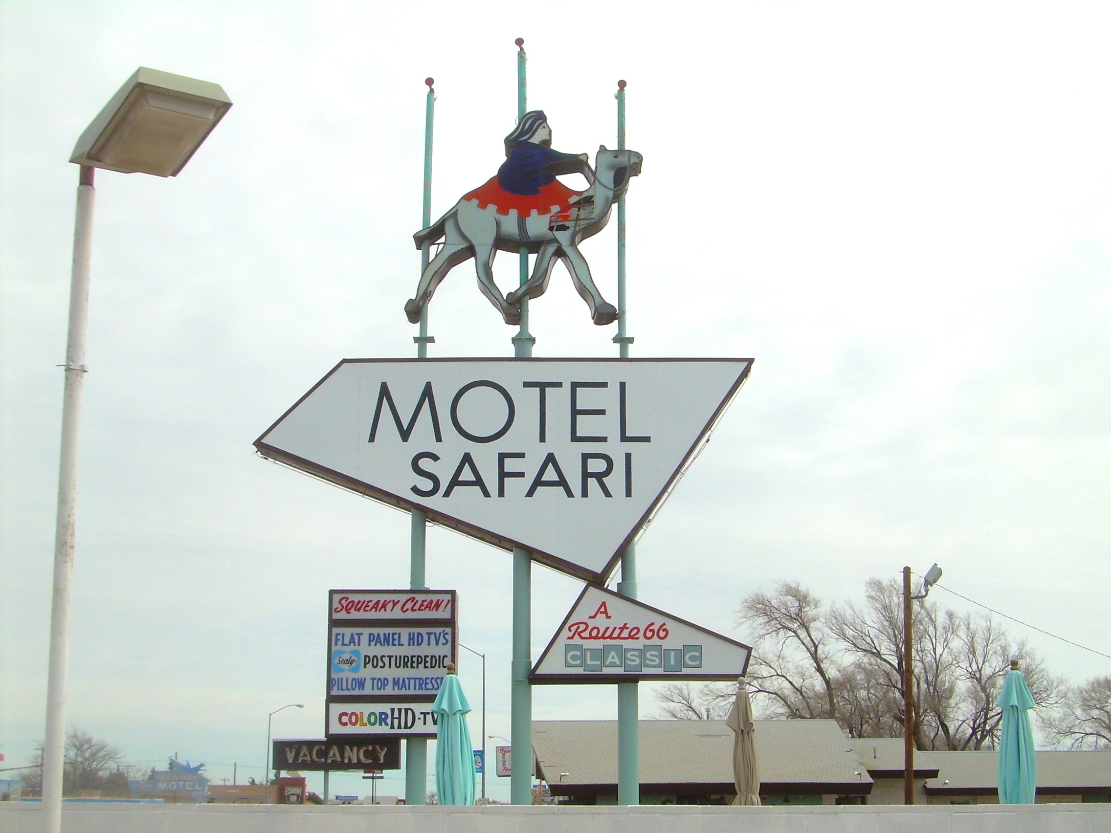 A large sign for Motel Safari featuring a camel rider statue and text indicating it is a Route 66 classic. The sign also advertises amenities such as flat panel