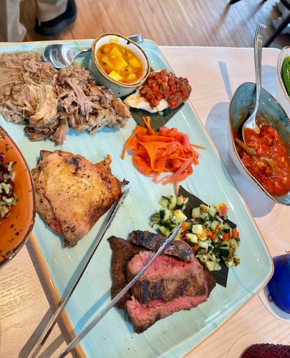 Family-style platter with sliced steak, chicken, pulled pork, vegetables, and sauces