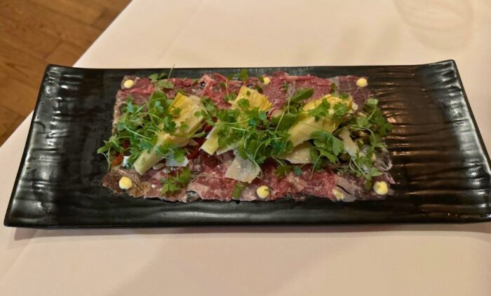 A plated dish of beef carpaccio garnished with herbs and artichokes, presented on a black rectangular plate.