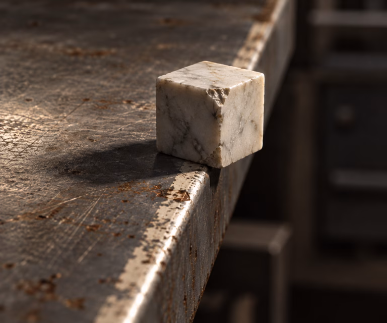 A chipped pale marble cube balanced with half its base overhanging the edge of a worn, rusted steel workbench under raking side light.