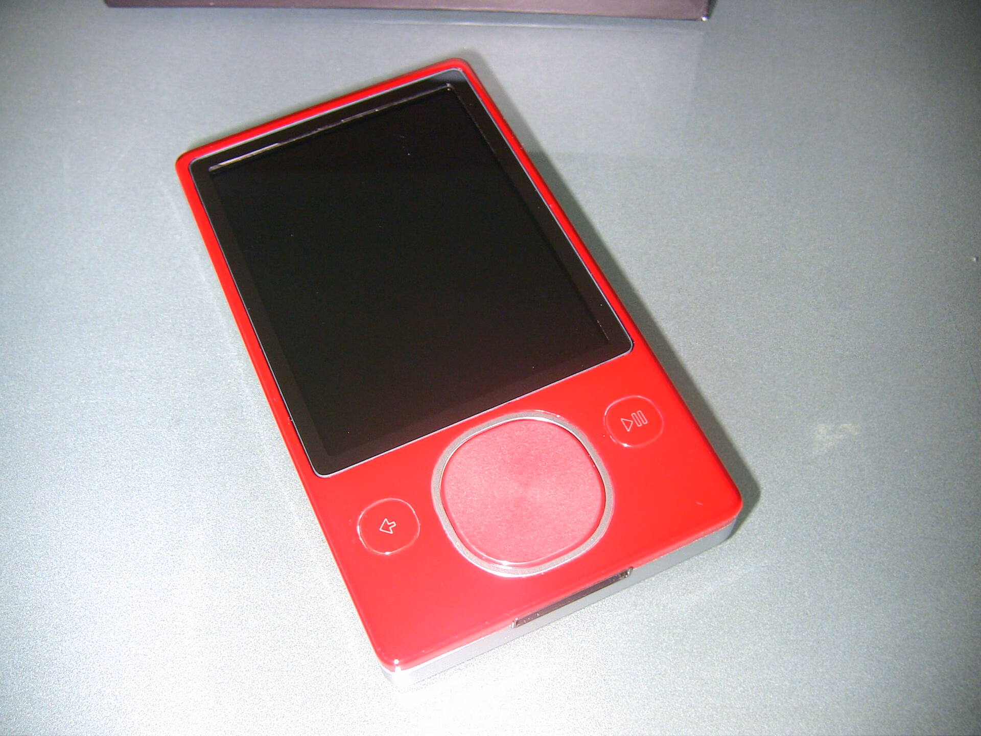 Red second-generation Microsoft Zune 80 with a touch-sensitive square pad and a blank screen, resting on a light green surface