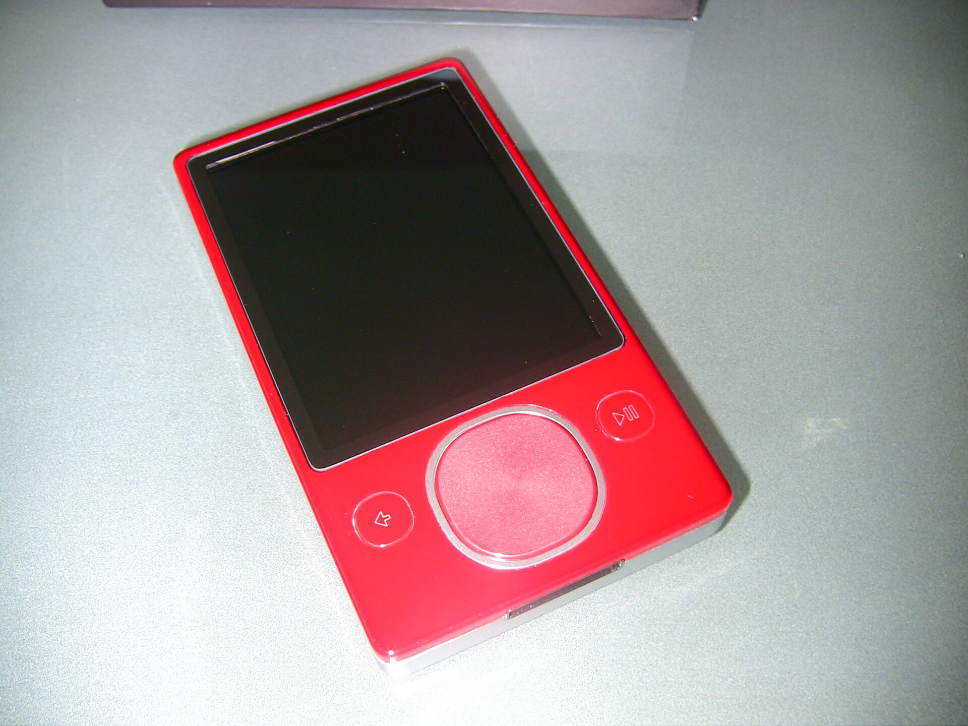 Red second-generation Microsoft Zune 80 with a touch-sensitive square pad and a blank screen, resting on a light green surface