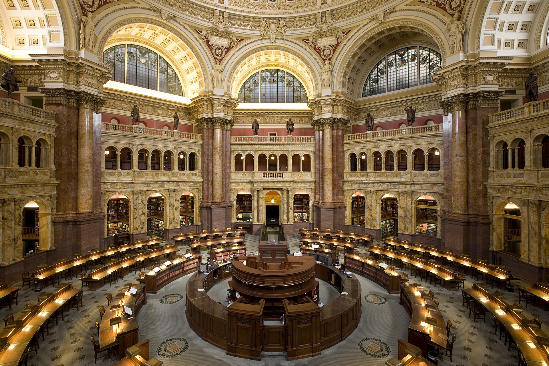 The octagonal Main Reading Room of the Library of Congress, with tiered bookshelves, a domed ceiling and rows of reading desks under warm light
