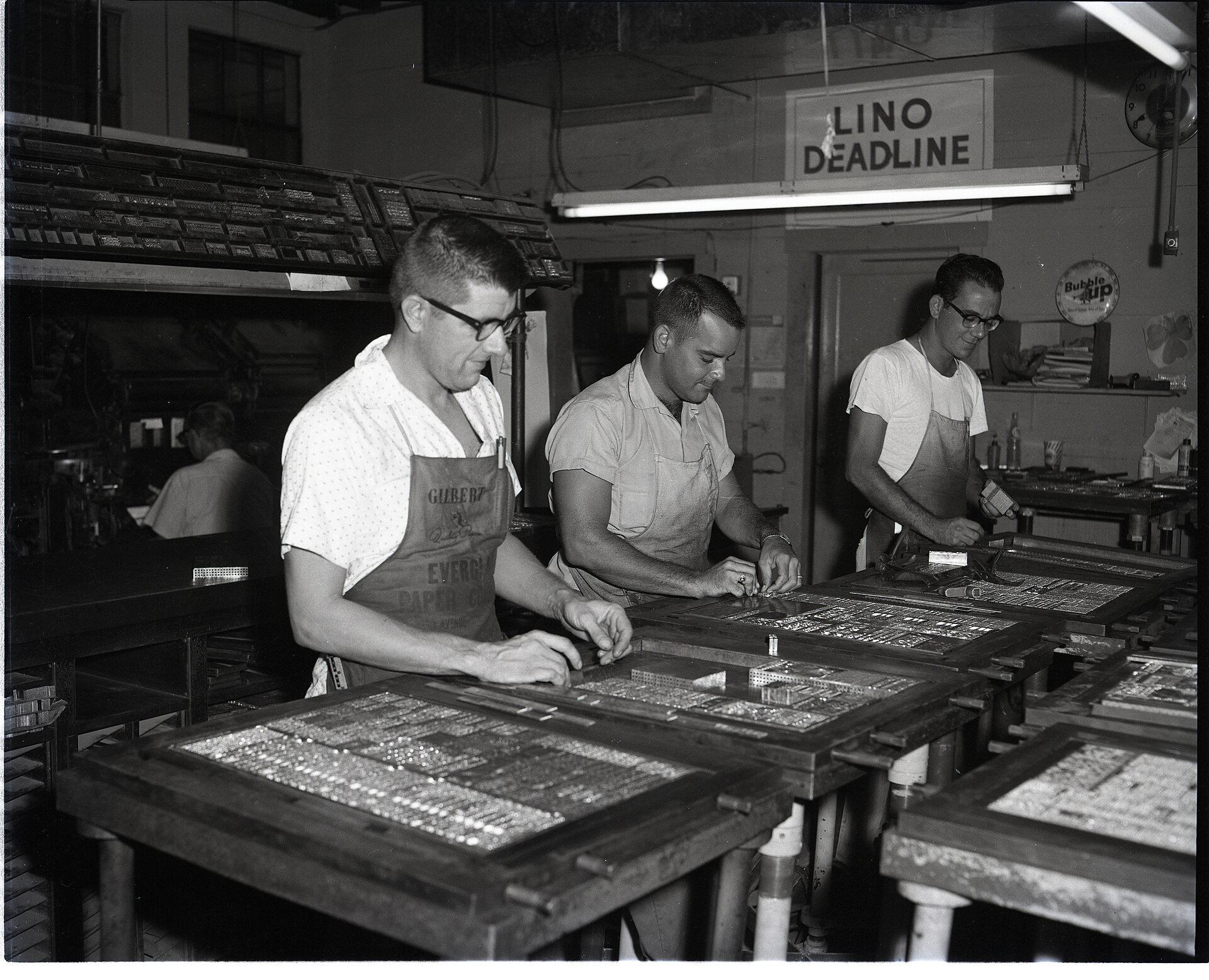 Three men assembling metal type for a newspaper page in a 1960s press room, with a Lino Deadline sign on the wall behind them